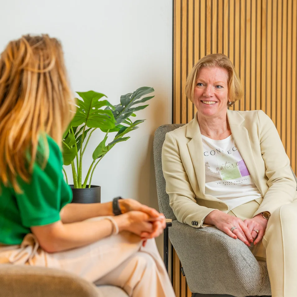 Susanne en Angelique zitten op twee stoelen. Angelique glimlacht naar Susanne